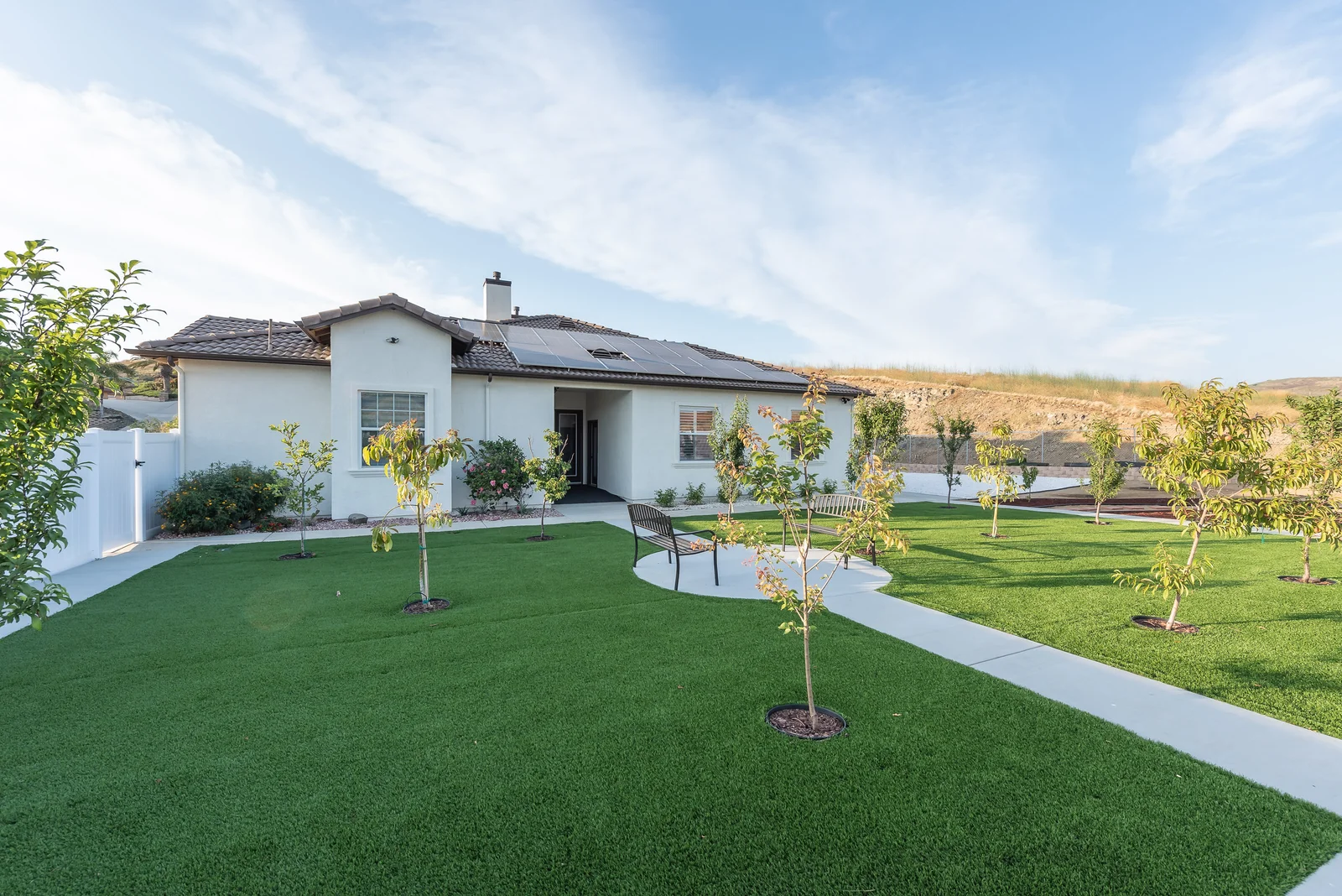 Backyard lawn with young fruit trees, a bench, and a paved walking path behind the Ohana Excellence home in Corona, California, with solar panels on the roof and hillside views beyond